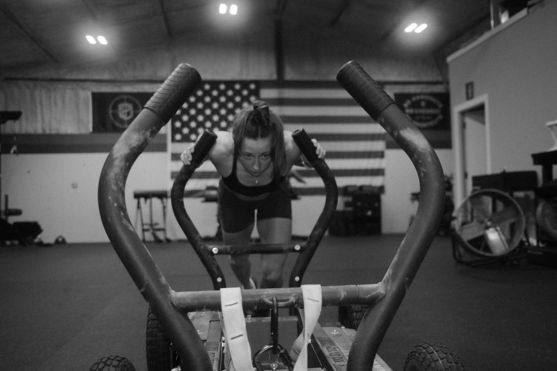 Black and white photograph of an athlete driving a weight sled head-on beneath an American flag